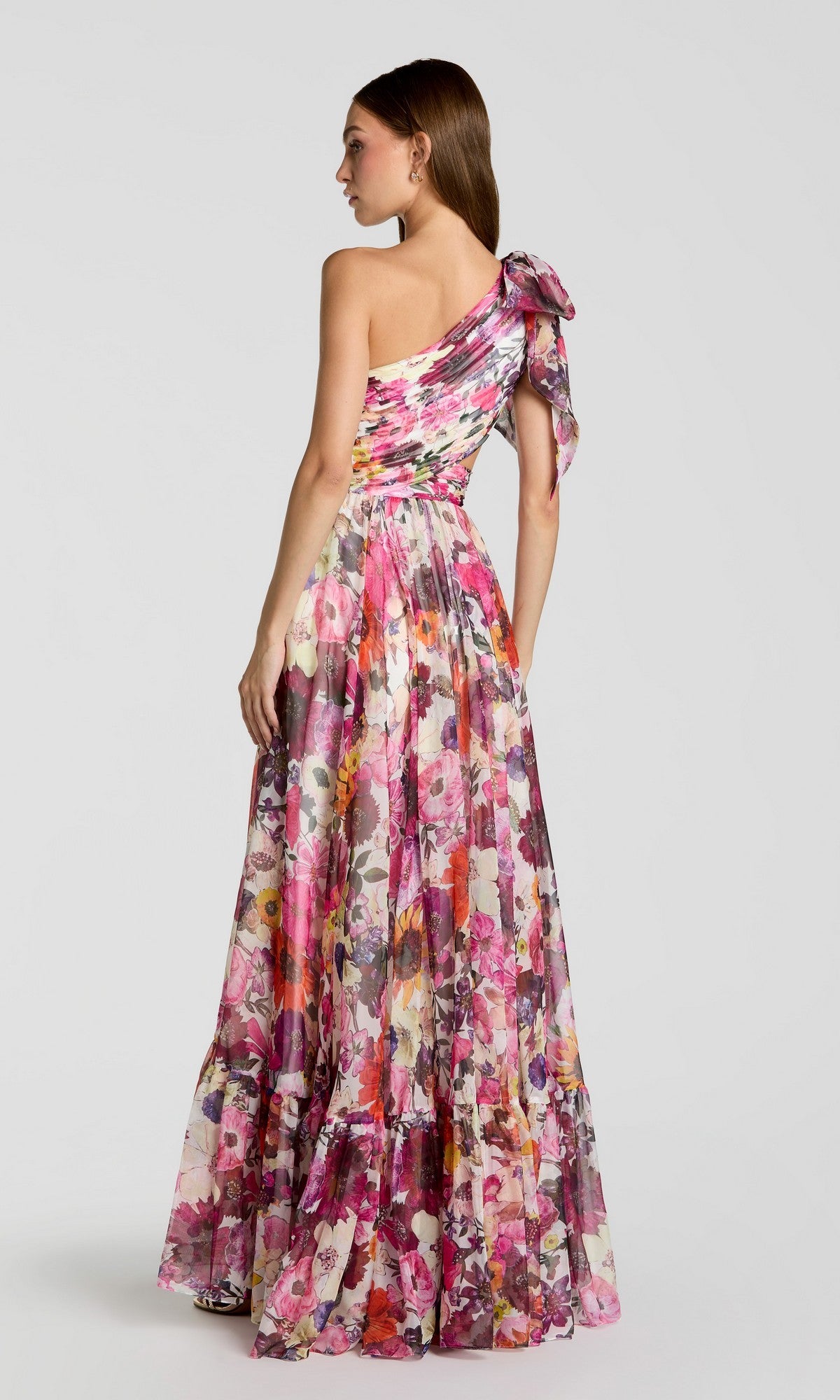 A woman faces away in the Mac Duggal 68553 long formal dress—an A-line, floor-length pink floral chiffon gown with a one-shoulder bow—set against a plain light background.